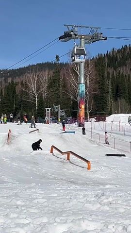 A sunny day at a ski resort features a snowboarder sliding down a low, orange rail in a terrain park. The snowboarder, dressed in all black, successfully navigates the rail and continues downhill.  Behind the snowboarder is a chairlift with empty gondolas, supported by a tall metal tower. The base of the tower has a colorful, decorative wrap. In the background, a hillside is covered with pine trees, with some bare birch trees closer to the ski area. Other skiers and snowboarders can be seen moving around the terrain park, and snow fences separate the terrain park from the other areas. The sky is clear and blue.