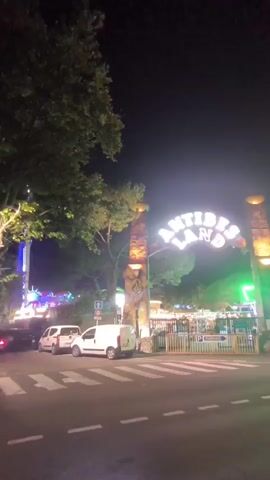 This is a video taken at night at an amusement park entrance. The entrance has a large archway with neon lights that spell out "ANTIBES LAND." To the left of the entrance, there is a tall amusement park ride with flashing lights. In front of the entrance, there is a crosswalk on a street with several parked cars, including two white vans. The ground is wet, suggesting it may have recently rained. Trees and foliage are visible around the entrance and the amusement park rides. The sky is dark, indicating nighttime.
