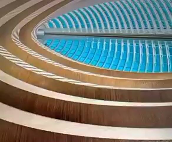 The video shows a close-up, slightly shaky view of a modern architectural interior. The focus is on a series of concentric, curved, wooden-paneled layers that create a spiral or tiered effect. Above these wooden layers, a bright blue, grid-like structure is visible, likely a ceiling or skylight made of glass panels. The overall impression is one of a spacious, contemporary building, possibly a public space like a museum, concert hall, or a large atrium. The lighting suggests daytime. There are no people or discernible actions occurring within the frame.