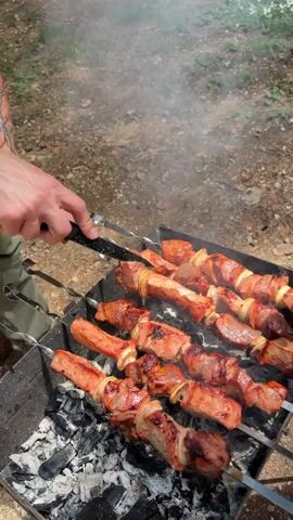 Two individuals are cooking meat on skewers over a charcoal grill in an outdoor setting. The first person, wearing green cargo pants and white sneakers, uses metal tongs to rotate and then remove cooked meat from a skewer. The second person, wearing green shorts and black slide sandals, holds a silver metal pot into which the cooked meat is transferred. Several skewers with grilled meat and onion segments are visible on the grill, emitting smoke. The ground is dirt, and green foliage is visible in the background.
