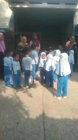 The video shows a group of young children, likely preschoolers, wearing matching blue pants and white shirts with some wearing hijabs. They are gathered outside what appears to be a school building with a blue wall and a banner displaying text. Adults, also wearing hijabs, are present, possibly teachers or parents. Some children are holding small plastic bags. The children seem to be lined up or milling about in a courtyard area. In the background, there are motorcycles parked, and the scene transitions to a street with more children and a residential area. The overall impression is of a school activity or departure.