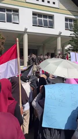 This video captures a protest taking place in front of a building, possibly a government or institutional building, in Kebuntanahterban, Indonesia. The time of day appears to be daytime, with bright sunlight casting shadows, suggesting fair weather.

The scene is populated by a crowd of people, many of whom are women wearing headscarves, indicating a cultural context common in Indonesia. The protesters are holding Indonesian flags, a mix of red and white, and several signs. One prominent sign, held by a person in the foreground, is a light blue poster with handwritten text in Indonesian. Visible text on this sign includes "KAMI BERSAMA PRABOWO" which translates to "We are with Prabowo." This strongly suggests the protest is in support of Prabowo, a prominent Indonesian political figure. Another smaller pink sign in the background has some visible text but is difficult to decipher fully.

The building in the background has a white facade with green trim and multiple windows. A covered walkway or balcony area on the second floor is visible, where several individuals in uniform, likely police or security personnel, are standing. They appear to be observing the crowd. Umbrellas, both white and pink, are being used by some protesters, suggesting the sun might be intense. The overall atmosphere seems to be one of a peaceful but determined demonstration. The presence of flags and signs, along with the gathering of people, clearly points to an organized political or social gathering.