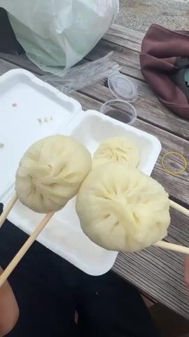 This media file captures a casual meal outdoors, likely at a roadside eatery given the visible "CU" convenience store signage and utility poles in the background. The setting appears to be Dongmyeong-myeon, South Korea. The weather seems overcast with a cloudy sky.

The primary focus is on a white styrofoam takeout container filled with several large, steamed dumplings. Two dumplings are held aloft on wooden skewers, presented to the camera. A small container of dipping sauce and a slice of bright yellow pickled radish are also visible within the takeout box. In the background, a brown bag and a white plastic bag rest on a weathered wooden picnic table, suggesting a relaxed, possibly impromptu meal.

The activity is the enjoyment of this food. The second half of the media shifts to a close-up of a young man wearing glasses and a green polo shirt. He is holding chopsticks with two dumplings, seemingly about to eat them. His expression is neutral, conveying a sense of simple pleasure or anticipation of the meal. The overall mood is one of everyday enjoyment and a simple, satisfying meal.

Visible text includes the "CU" logo on a purple sign, a common South Korean convenience store chain. There is also some Korean text on a sign in the background, but it is too small and blurry to be legible.