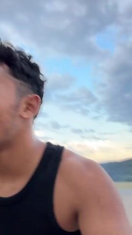 This media file features a close-up shot of a person from the shoulders up, seen from the side. The individual has dark, curly hair with a faded haircut on the sides and is wearing a black tank top. Their skin appears tanned, and some sweat is visible on their neck and shoulder, suggesting physical activity. The background is dominated by a cloudy sky with patches of blue, indicating a partly cloudy day. The lighting suggests it is either early morning or late afternoon, given the soft, diffused light.

A hint of a wooden surface is visible at the bottom left of the frame, implying the person might be on a deck or a wooden platform. In the distance, a hazy outline of hills or mountains can be seen. The overall mood is candid and natural, with no overt display of emotion. The media appears to be a still frame taken from a video, as indicated by the timestamp "00:00" to "00:04" at the beginning of the file. There is no visible text in the media. The location context provided is Pavel Banya, Bulgaria.