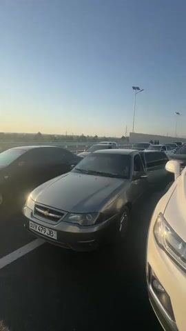 The video shows a traffic jam on a clear day, with many cars lined up on a road. The camera is positioned low to the ground, looking up at the sky and across the traffic. In the foreground, a white SUV is parked, and a blue bottle of water is visible on its hood. To the left of the SUV is a gray sedan, and further back, a long black limousine is visible. Other cars, including SUVs and sedans, are seen in the background, stretching as far as the eye can see. People are visible in some of the cars and standing outside of them. The environment appears to be urban or suburban, with buildings and streetlights visible in the distance. The sun is shining, casting long shadows on the road.