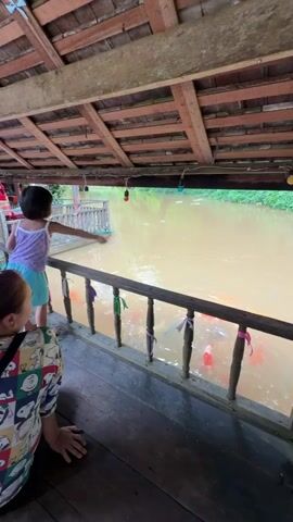 A child feeds fish from a wooden deck over a muddy canal, observed by an adult sitting on the floor.