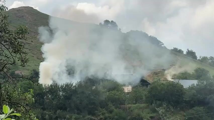 The video shows a rural landscape with a hill in the background and trees in the foreground. There are several structures that appear to be houses or cabins visible through the trees. A large amount of white smoke is billowing from one of the structures on the left side of the frame. Another smaller plume of smoke is rising from a structure on the right side of the frame. The sky is cloudy. The smoke suggests that there may be fires burning at the structures, or possibly some kind of controlled burning activity.