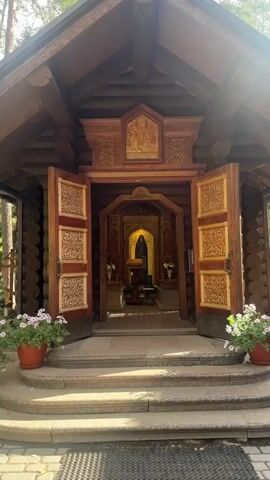 The video shows the entrance to a small wooden Orthodox chapel. The chapel has a double wooden door with intricate carvings on both sides. The door is open, revealing an altar with an icon of a bearded man in religious attire. The altar is decorated with flowers and candles. The exterior of the chapel is made of logs, and there are potted plants on either side of the entrance steps. Sunlight streams in from the left side, creating a bright spot on the chapel's roof. The video zooms in on the altar and the icon.