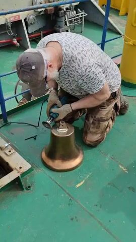 A man is kneeling on a green deck and using a power tool to clean a bronze-colored bell. The man is wearing a hat, a white patterned shirt, and camouflage pants. He has gloves on and appears to be focused on his task. The bell sits on the deck in front of him. The background includes metal structures and railings painted in blue and yellow. The scene suggests the man is likely on a boat or ship.