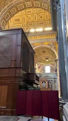 The video opens with an upward-angled shot inside a large, ornate building, likely a church or cathedral. A dark wooden structure, possibly an organ or confessional, stands prominently in the foreground on the left. To its right, red fabric screens off an area. The floor is tiled with geometric patterns. The ceiling, vaulted and grand, is adorned with intricate gold and white patterns, and a large golden dome with paintings is visible further back. As the camera pans to the left, a marble altar or monument with a white statue comes into view. The camera continues to pan upward and left, revealing more of the richly decorated ceiling and domes, featuring detailed frescoes and golden embellishments. Natural light streams in from windows high up. The video ends with a view of the multiple domes and vaulted ceilings, emphasizing the opulent architecture.