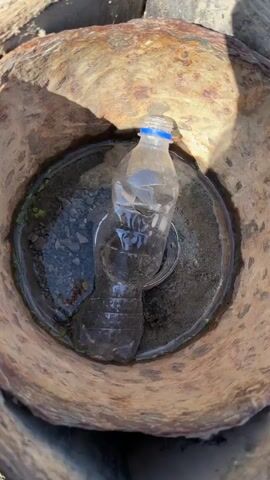 A person is emptying a clear plastic water bottle into a rusted metal container filled with water. The water in the container is slightly murky and contains small pebbles and bits of vegetation. The container itself has a weathered, brownish-orange surface with dark patches of rust.  A circular metal piece lies submerged near the center of the water.  The person's hand, partially gloved with a black and yellow sleeve visible, is seen holding and tilting the bottle to pour out its contents.  The action takes place outdoors under natural light.