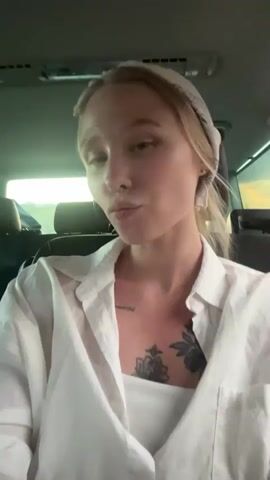 A young woman with blonde hair, wearing a white button-down shirt and a white bandana, is seated in the back of a vehicle. She has a tattoo of a flower on her chest and a nose ring. She is looking at the camera and speaking. The interior of the vehicle is visible, with headrests and ceiling lights. Through the windows, a bright, possibly sunny sky can be seen. The scene appears to be taking place during the daytime. The woman's expression suggests she is engaged in conversation or singing. The visible text is in Russian and reads: "Ты такая красивая, я перестаю дышать" which translates to "You are so beautiful, I stop breathing." The location context is Mišići, Montenegro.