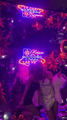 The video shows two young women kissing in front of a "Welcome to Fabulous Las Vegas" neon sign. They are dressed in party attire, one in a sequined black dress and the other in a white dress with a fluffy white jacket. The background includes party decorations such as star-shaped balloons and disco balls. A few other people are visible in the background, dancing and talking. The overall atmosphere is that of a lively party or celebration with purple and pink neon lighting.