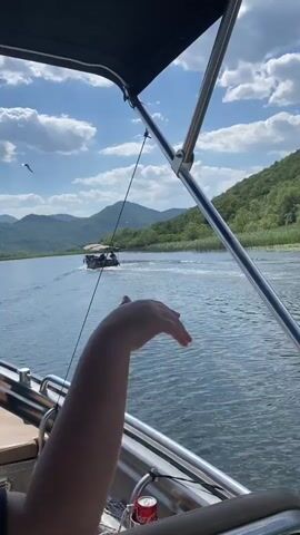 The video shows a bright, sunny day on the water. Two boats are visible. In the foreground, the camera is positioned on a boat, looking out at the water. A person's hand is resting on the edge of the boat in the bottom right corner. In the middle ground, a larger, wooden boat with a canopy is traveling away from the camera. This boat is carrying several passengers, some of whom appear to be waving. The water is calm with gentle ripples. In the background, lush green hills rise steeply from the edge of the water, with some trees and reeds along the shoreline. The sky is blue with fluffy white clouds. The overall impression is one of a pleasant boat trip on a warm day in a scenic, natural environment. The location cues of green hills and a lake suggest a natural landscape, and the presence of a tourist boat filled with passengers implies a recreational or sightseeing activity.