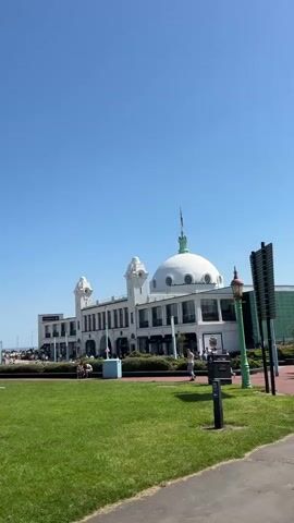 The video showcases a sunny day at a park with a large green lawn. In the background, a white building with a distinctive dome can be seen. People are scattered across the lawn, some sitting and relaxing, others walking.  A stroller is parked near a family sitting on the grass. A monument and the sea are visible further in the background. The sky is clear and blue. The scene appears peaceful and relaxed.