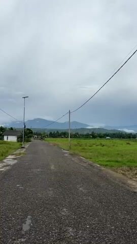 The video shows a view of a road that goes into the distance, flanked by green fields on both sides. In the background, there are mountains with some cloud cover. There are power lines and poles along the road, and a streetlight on the left side of the road. The sky is overcast with clouds.