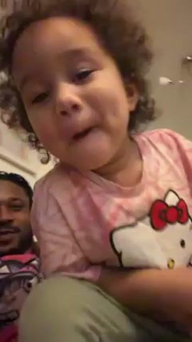 A close-up video shows a young child with curly hair wearing a pink Hello Kitty shirt, sitting in front of a man with dreadlocks. The child opens their mouth wide and then closes it in a playful manner, smiling. They look directly at the camera. The man is sitting behind the child, partially visible in the background. The background includes a wall, a framed picture, and a light fixture. Music is playing in the background.