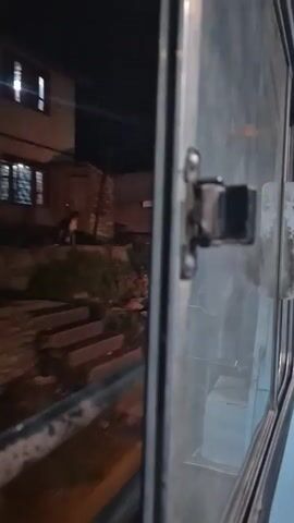 The video appears to be filmed from inside a moving vehicle, possibly a train or bus, looking out a window. The window is dirty and has a metal grate along the bottom. The exterior view shows a street at night. There are buildings with shuttered storefronts, one with a yellow facade and an advertisement that includes the word "KAMACH." Greenery, possibly plants or bushes, is visible along the edge of the view. Lights from the buildings and street provide illumination. The camera is mostly focused on the window and its latch, with the outside view slightly blurred due to motion.