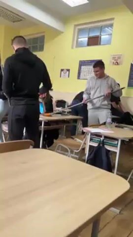 The video shows the interior of a classroom. Several students are standing around desks, some holding long, metal poles. One student, wearing a gray hoodie, is holding a pole and smiling, while another student in a black sweatshirt is manipulating a cleaning device at the end of a pole, possibly a vacuum cleaner attachment. The classroom has yellow walls, several windows, and posters on the walls. Desks are wooden with metal frames. The overall atmosphere appears to be informal and somewhat chaotic.