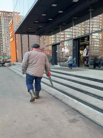 The video depicts an urban street scene outside a modern building with large, reflective glass windows. The primary focus is on a man, viewed from behind, walking along a concrete sidewalk towards the building's entrance, which is accessed by a set of wide concrete steps.

The man is dressed in a light-colored, possibly grey or beige, jacket, blue jeans, and dark green rubber boots. He wears a dark cap on his head. In his right hand, he carries a handsaw with a distinct red handle, letting it swing loosely by his side as he walks.

In the background and reflected in the glass doors of the building, other people are visible. A woman is seen pushing a baby stroller up the steps towards the entrance. A small child is also visible on the steps, possibly running. Further in the background, another person with a stroller can be seen on the left side of the frame.

The building itself has a contemporary design with dark frames around the large glass panels and a dark, possibly metal, overhang above the entrance. The reflections in the glass show other buildings and the general surroundings. The weather appears to be daylight, possibly overcast, with soft lighting. The audio includes a distinct squeaking sound, likely from a stroller wheel, along with general ambient noise. No clear, legible text is visible on any signs or the building.