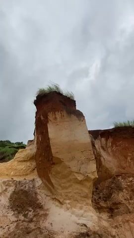 A video shows a rocky outcrop, likely a cliff face or eroded hillside. The rock layers are distinct, with a top layer of dark reddish-brown soil and vegetation, followed by lighter tan and yellow rock layers beneath. The rock surface is rough and appears to be a natural formation, possibly affected by erosion. The sky is overcast with gray clouds, suggesting a dull or possibly rainy day. The lighting is soft and diffused. The camera movement is subtle, with slight zooms and pans that focus on the texture and geological layers of the rock formation. There are no people or animals visible in the video. The environment appears to be natural and undeveloped.