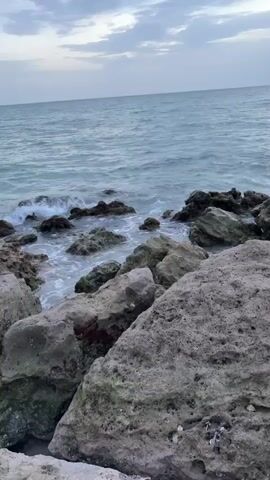 The video shows a rocky shoreline with the ocean in the background. The water is a murky blue-green and appears to be somewhat choppy, with waves breaking against the rocks. The sky is overcast with gray clouds. The rocks in the foreground are large and rough, with some appearing to be covered in lichen or algae. The overall impression is of a rugged and somewhat desolate coastal scene.