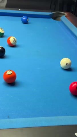 The video shows a billiard table with several billiard balls scattered across its blue surface. A cue stick is visible, positioned as if about to strike one of the balls. The billiard balls are various colors and numbered, including green, orange, blue, black, and red. The table has a blue felt surface and dark-colored rails. There's a faint glimpse of people standing in the background, though they are out of focus. A successful shot results in the striped ball bouncing into the side rail.