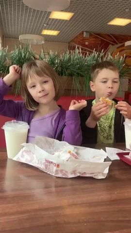 Two young children, a girl and a boy, are seated at a brown table with red booth seating in what appears to be a fast-food restaurant. The girl, on the left, has short light brown hair and is wearing a purple jacket over a lighter purple top. She makes various playful hand gestures, smiles at the camera, and has a white drink in a clear plastic cup beside her. The boy, on the right, is wearing a black jacket over a green top. He is eating a small, round food item, possibly a burger, and at one point holds it up to partially cover his face. Empty food wrappers are on the table in front of both children. Artificial plants decorate the background.