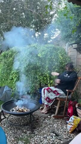 A woman sits in a rocking chair, waving her hands to deflect smoke rising from a fire pit, in a backyard setting. The woman is wearing a dark sweatshirt and patterned pants, and her bare feet are visible. The fire pit, made of metal, contains burning pieces of wood and is surrounded by gravel. Behind the woman is a lush green weeping willow tree and a wooden structure, likely a shed. Bags of chips and a bottle are visible next to the chair. The scene is shot outdoors during daylight.