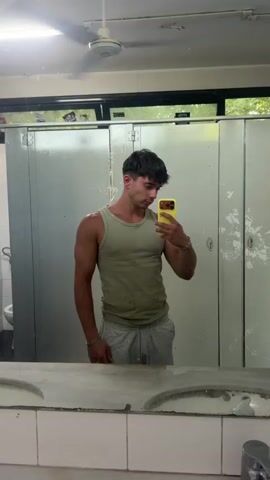 A young man with dark hair, wearing a light green tank top and grey sweatpants, stands in front of a mirror and takes a selfie with his yellow-cased smartphone. The man is muscular and appears to be in a locker room or bathroom, as evidenced by the stalls, sink, and fan visible in the background. The lighting is bright and overhead.