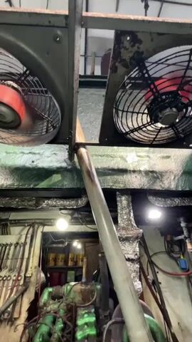 The video pans across a mechanical room or engine room. Two large industrial fans are visible at the top, one on the left with red accents and the other on the right. Below the fans, a large, green engine dominates the center of the frame. Various pipes, hoses, and wiring are connected to the engine. To the left of the engine, a wall is lined with tools, including wrenches and other mechanical instruments. In the background, yellow containers are stacked. The lighting appears to be artificial, with several light fixtures illuminating the space. The environment seems somewhat industrial and utilitarian, with visible insulation and metal structures.