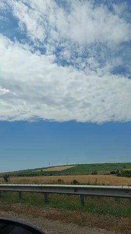 This is a horizontal, first-person perspective video of a car driving through a rural landscape on a sunny day. The sky is blue with scattered white clouds. On the left, there is a field of dry grass and vegetation. A metal guardrail is visible in the foreground. On the right, there is a sign with a yellow background and a red arrow pointing to the right. Beyond the guardrail, there are fields of golden crops, green fields, and a few distant windmills. Trees are visible on the right side of the frame. The video captures the motion of the car, indicated by the changing scenery and the blur of the passing landscape.
