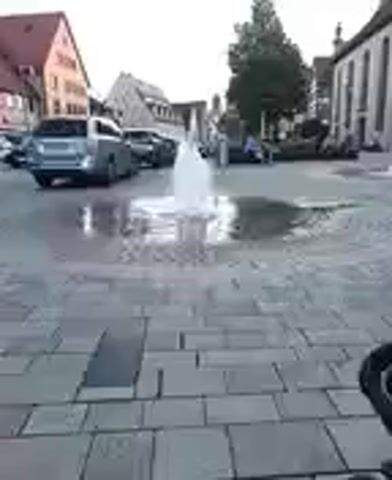 The media file shows a public square in Altdorf bei Nürnberg, Germany. The scene is paved with cobblestones and features a water fountain in the center, creating a refreshing spray. In the background, buildings with traditional German architecture are visible, including houses with gabled roofs and a larger building with a tower, suggesting a historical town center. Several cars are parked on the street, and some people can be seen sitting or walking around the fountain, enjoying the atmosphere. The lighting suggests it is late afternoon or early evening, with soft, diffused light. The overall mood appears calm and relaxed, with people enjoying a pleasant day outdoors. There is no visible text in the media file.