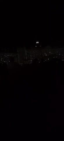 The video shows a dark outdoor scene, possibly a stadium or concert venue, at night. The foreground is almost entirely black, with silhouettes of people visible. In the background, there is a brightly lit area, suggesting a stage or performance area with many lights. A tall, slender structure, possibly a tower or building with lights, is visible in the upper center of the frame. The overall impression is of a large gathering of people at an event taking place after dark.