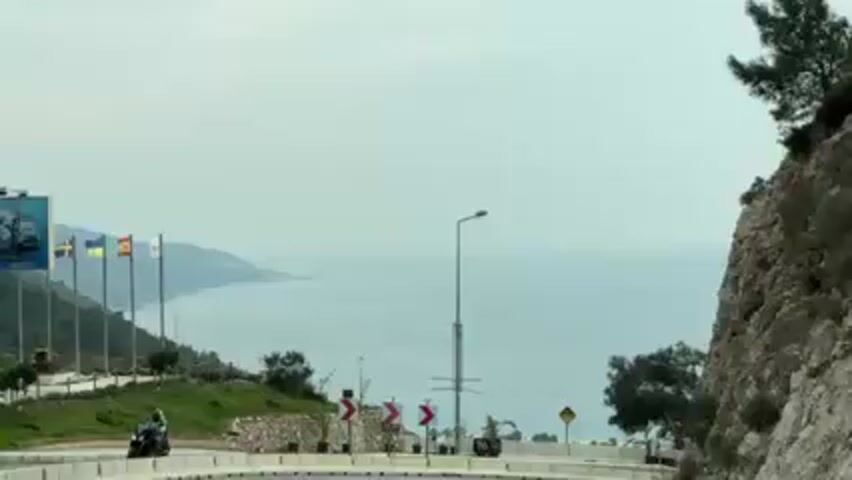 The video shows a coastal view from a road. Along the roadside, there are several national flags on poles, including those of Sweden, Spain, and Ukraine. A white van with "TAXI" written on the side is parked next to a tree. Road signs are also visible. The horizon reveals a coastline with mountains in the distance and the sea under a cloudy sky. A motorcycle passes by in the first few frames.
