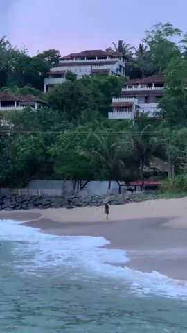 This media file captures a serene beach scene at dusk, likely in Batawala, Sri Lanka. The foreground is dominated by the ocean, with gentle waves lapping at the shore, their white foam creating a beautiful contrast against the darker water. A sandy beach stretches across the middle ground, where a lone figure, a person wearing a dark swimsuit, is seen standing near the water's edge, looking out towards the sea. Their solitary presence suggests a moment of quiet contemplation or enjoyment of the peaceful surroundings.

Behind the beach, a line of dark rocks and a white picket fence delineate the property from the lush, green hillside. The hillside itself is densely covered with tropical trees and palm trees, their vibrant foliage creating a rich tapestry of green. Perched atop this verdant slope are several white, multi-story buildings with red-tiled roofs. These appear to be villas or resort accommodations, their architecture hinting at a tropical or Mediterranean influence. The buildings are nestled amongst the trees, blending harmoniously with the natural landscape.

The time of day is clearly evening, as indicated by the soft, diffused light and the purplish hue of the sky, suggesting either sunrise or sunset. The weather appears calm and pleasant, with no strong winds or harsh sunlight. The overall atmosphere is one of tranquility and natural beauty, evoking a sense of escape and relaxation. The media file contains no visible text.