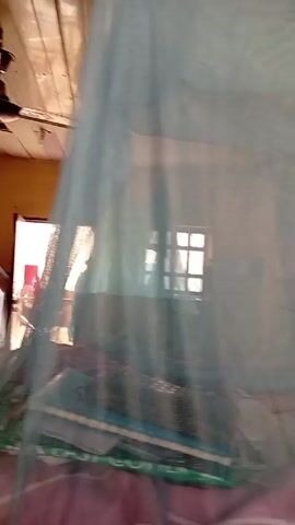 The video shows a modestly furnished room with two beds under mosquito nets. The walls are painted a light yellow and the ceiling is wooden. A ceiling fan is visible.  There are windows and a doorway in the background.  The room appears to be lived in, with various items scattered around, including a bucket and bags. No people are visible.