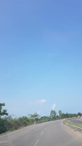 This video captures a scenic drive along a highway, likely in Mudaliyarkuppam, India. The primary subjects are the road itself and the surrounding natural environment, dominated by a vast, clear blue sky.

The scene depicts a road that curves gently to the right, bordered by lush green vegetation and trees. The road surface appears to be asphalt, with clear white lane markings visible. There are some signs along the roadside, but the text is not discernible.

Objects visible include occasional vehicles on the road, including what appears to be a motorcycle and possibly a car further in the distance. There are also what look like guardrails and concrete barriers along the side of the road. The vegetation suggests a warm climate, with leafy trees and some dry brush.

The activity taking place is vehicular travel along the highway. The camera seems to be mounted on a moving vehicle, providing a first-person perspective of the journey.

Notable details include the bright, clear blue sky, indicating it is daytime with good weather, likely a sunny day. The time of day appears to be late morning or early afternoon, given the bright light and the quality of the shadows (though shadows are not a prominent feature in this wide-angle shot). There are a few wispy clouds scattered across the sky. The overall mood conveyed is one of peaceful travel and appreciation for the natural landscape.

The location context of Mudaliyarkuppam, India, suggests a possibly coastal or rural setting, which aligns with the visual cues of abundant greenery and open sky.