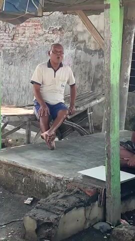 A middle-aged man sits on a low platform with his legs extended. He wears a light-colored polo shirt and blue shorts. The man is looking upwards and appears to be speaking or singing. He is in an outdoor setting under a simple, open-air shelter with a corrugated roof. The shelter is supported by wooden posts, one of which is painted green. Behind the man, a brick wall is visible. The platform is made of concrete and there are some pieces of wood and debris on the ground in front of it. To the right of the frame, another person's legs are visible.