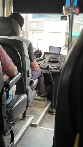 The video shows the interior of a bus from a passenger's perspective, looking towards the front. A male bus driver, seen from behind, is seated in the driver's seat. He is wearing a dark t-shirt and dark pants, with brown shoes. His left hand is on the steering wheel, and his right hand is near the gear shift. A digital screen is mounted on the dashboard in front of him. Various buttons and controls are visible on the dashboard. There are also passengers seated in the front section of the bus, with the back of a passenger in a red shirt visible on the left and the side of another passenger on the right. The bus appears to be in motion, as indicated by the blurred scenery outside the windshield, which includes buildings, trees, and traffic lights. The overall environment suggests a daytime scene on a city street.