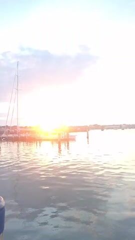 The video shows a sunset over a body of water. In the foreground, there is a dock with a sailboat and several other boats. The water is calm, and the sky is filled with clouds. The sun is low on the horizon, and its rays are reflecting off the water.