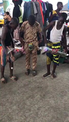 Three young Black boys are standing in a line in what appears to be an outdoor market. They are all looking down and engaged in some activity. The boy on the left is wearing a black tank top and colorful shorts. He has a small, dark-colored object in his hands and is focused on it. The middle boy is wearing a tan and brown patterned outfit, including a long-sleeved shirt and pants. He is also looking down at something held between his hands. The boy on the right is wearing a white tank top and a patterned blue, yellow, and black wrap skirt. He is holding a colorful object with both hands. All three boys are wearing sandals. In the background, there are racks of clothing for sale, and other people are visible. The ground is paved with concrete.