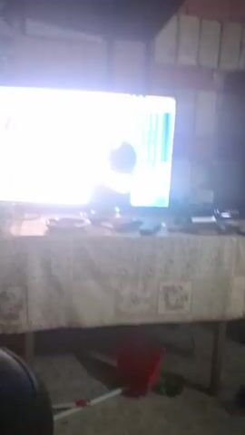 The video shows a TV screen displaying various images of beaches, including palm trees, blue water, and people. A table below the TV holds several items, including what appears to be a cup, a plate, and a remote control. The room itself appears to be a simple living space, possibly in a rustic setting, with visible brickwork. The camera angle shifts slightly during the video. There is music playing in the background.
