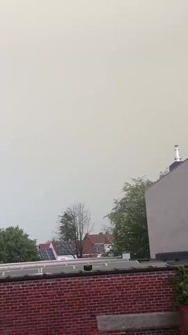 The video captures a brief moment of a thunderstorm from a distance. The scene shows a grey, overcast sky. In the foreground, the tops of trees are visible, with some bare branches suggesting the season or the state of the trees. Behind the trees, the roofs of houses can be seen, some equipped with solar panels. The most prominent event is a flash of lightning that briefly illuminates the sky with a bright white light, followed by a faint purple hue, indicating the intensity of the electrical discharge. The lighting is otherwise dim, consistent with stormy weather. There are no people visible in the video.