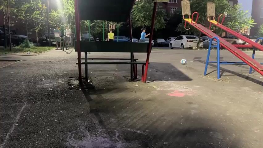 The video captures a playground scene at dusk, illuminated by streetlights. In the foreground, a gravel path shows patches of chalk drawings, with shadows cast from overhead structures. To the left, a covered picnic table with benches is partially visible, with a few people standing around it. In the center of the frame, a soccer ball rests on the ground near a young boy in a blue shirt who appears to be playing soccer. To the right, a red seesaw with blue supports is present, with a child actively swinging on one side. Further in the background, several parked cars are visible, and residential buildings loom beyond the playground. The overall atmosphere suggests a casual evening gathering of children and possibly adults in a neighborhood playground.