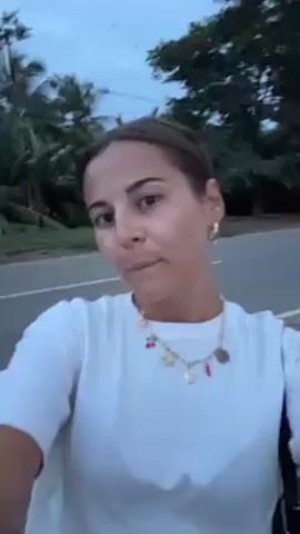 A woman takes a selfie video outside. She wears a white shirt, a short denim skirt, and black flip-flops. She has dark hair pulled back into a bun and is wearing a necklace with charms. She smiles at the camera. The background is a paved area.