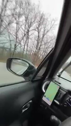 A driver is traveling on a rainy road. The driver, as visible in the sun visor mirror, has short dark hair and is recording with an iPhone. The car is a dark color on the inside, the dashboard featuring a speedometer, navigation on a phone mount, and wipers. Outside, trees are visible in a forest on either side of a wet, narrow road.