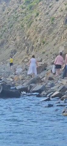 The video shows a rocky shoreline with a steep, tan-colored cliff face in the background. Several people are walking along the shore. In the foreground, the blue water of the sea is visible with small waves. From left to right, a person in a yellow shirt and dark shorts is walking away from the camera. Next, a person in a white dress is walking away from the camera. Further to the right, a person in a pink shirt and dark shorts is walking away from the camera, carrying a white bag. Next to them, a person in a light-colored shirt and blue shorts is walking away from the camera, carrying a dark bag and a blue bag. The ground is covered with numerous rocks of varying sizes. Some sparse green vegetation is visible on the cliff face.