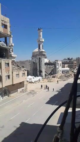 This media file depicts a scene of extensive destruction in an urban area, identified as Khan Yunis in the Palestinian Territory. The central focus is a heavily damaged mosque with a prominent, but also damaged, minaret still standing. Its dome is partially missing, and large sections of the mosque's structure appear to have collapsed into rubble.

Surrounding the mosque are multi-story buildings, many of which are severely damaged or reduced to partial ruins. Walls are blown out, interiors exposed, and debris is scattered extensively across the scene, particularly around the base of the mosque and along the sides of the paved street. The street itself appears largely intact, albeit with some scattered rubble.

Several people, including adults and at least one child, are seen walking along the street. They are moving through the devastated environment, suggesting an ongoing presence of residents despite the destruction. One cluster of individuals is visible near the entrance of the damaged mosque.

The video is recorded on a bright, sunny day with a clear blue sky, casting distinct shadows, indicating a mid-morning or early afternoon time. The weather appears calm. The perspective is from an elevated position, possibly a balcony or higher floor of a building, looking down onto the street and the ruined landscape. A dark metal railing is visible in the foreground, partially obstructing the view.

Visible text on signs is mostly in Arabic. On a damaged building to the left, a sign contains partially legible text, including "CAT." On a sign in the lower right, the word "أفراح" (Afrah, meaning "joys" or "weddings") is somewhat discernible, though the full context of the sign is unclear. The overall scene conveys a profound sense of devastation resulting from conflict, yet life continues as people navigate the altered landscape.