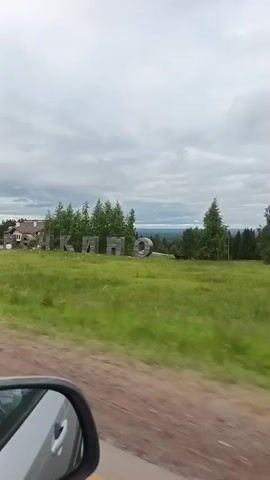 A video from the perspective of a vehicle shows a large, stylized sign with Russian letters on a grassy hill. The camera pans from left to right, revealing the letters "ЧКАЛО", which is transliterated to "CHKALO". In the background, there's a forest, and a white house with a brown roof is visible to the left of the sign. The sky is overcast. A rearview mirror is visible in the bottom left corner of the frame, reflecting the interior of the vehicle.